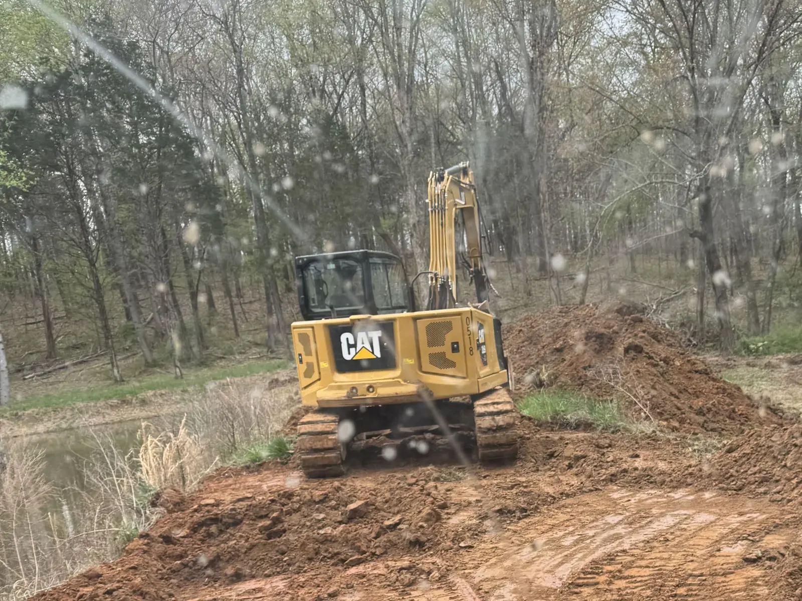 Excavator on the regraded dam top with the woods behind, seen from the loader cab