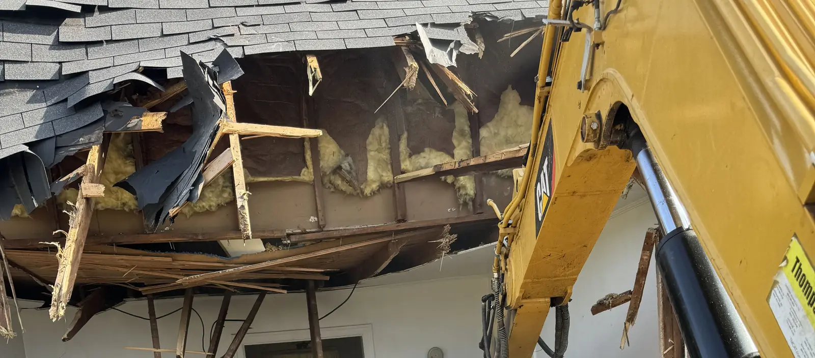Excavator boom through the torn-open roof of a house during demolition, insulation and broken rafters hanging