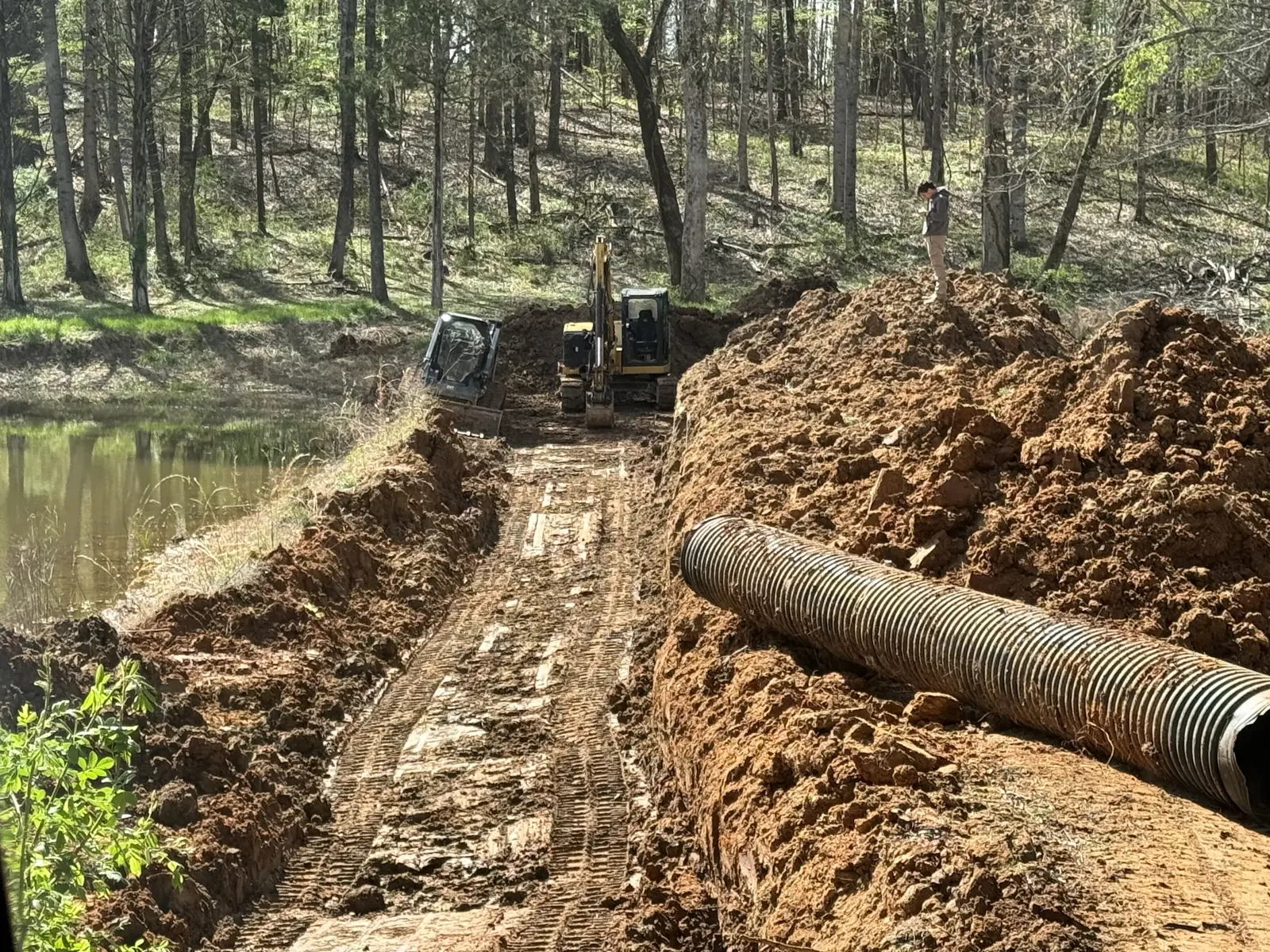 Corrugated pipe laid beside the trench cut through a pond dam, the excavator and the track loader at the far end