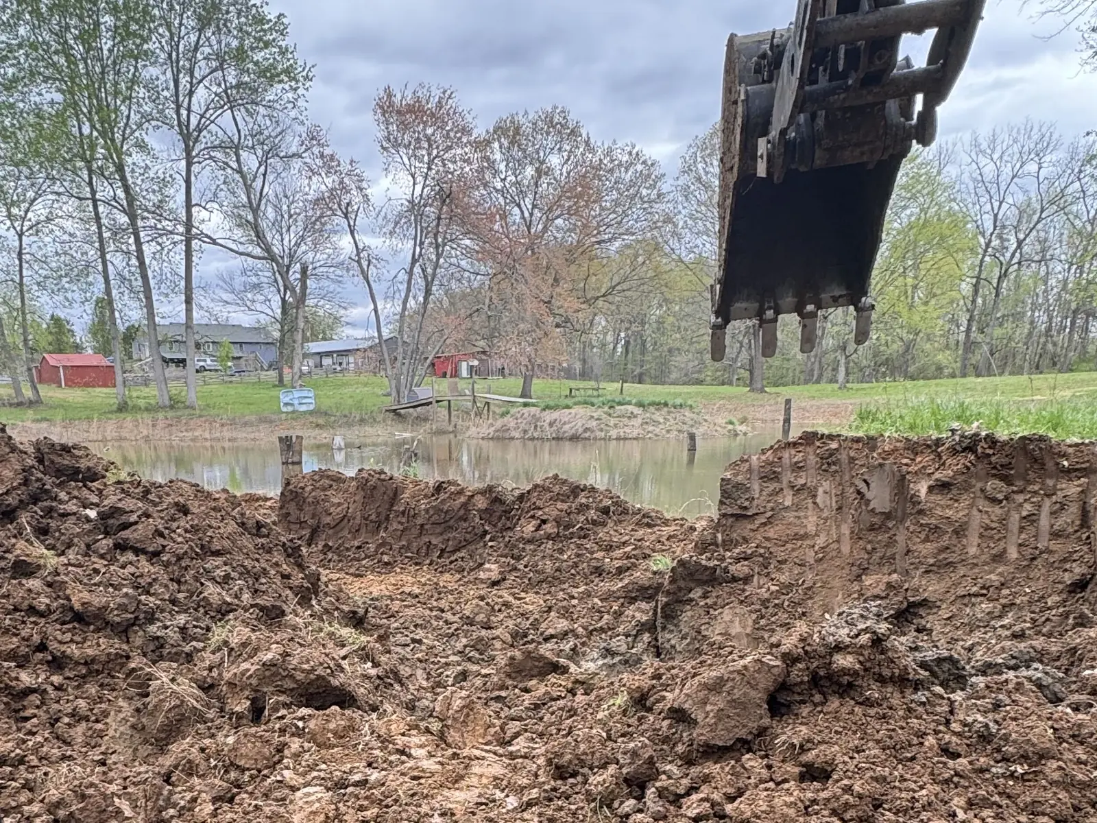 Excavator bucket cutting a trench down through a pond dam, seen from the cab with the pond beyond