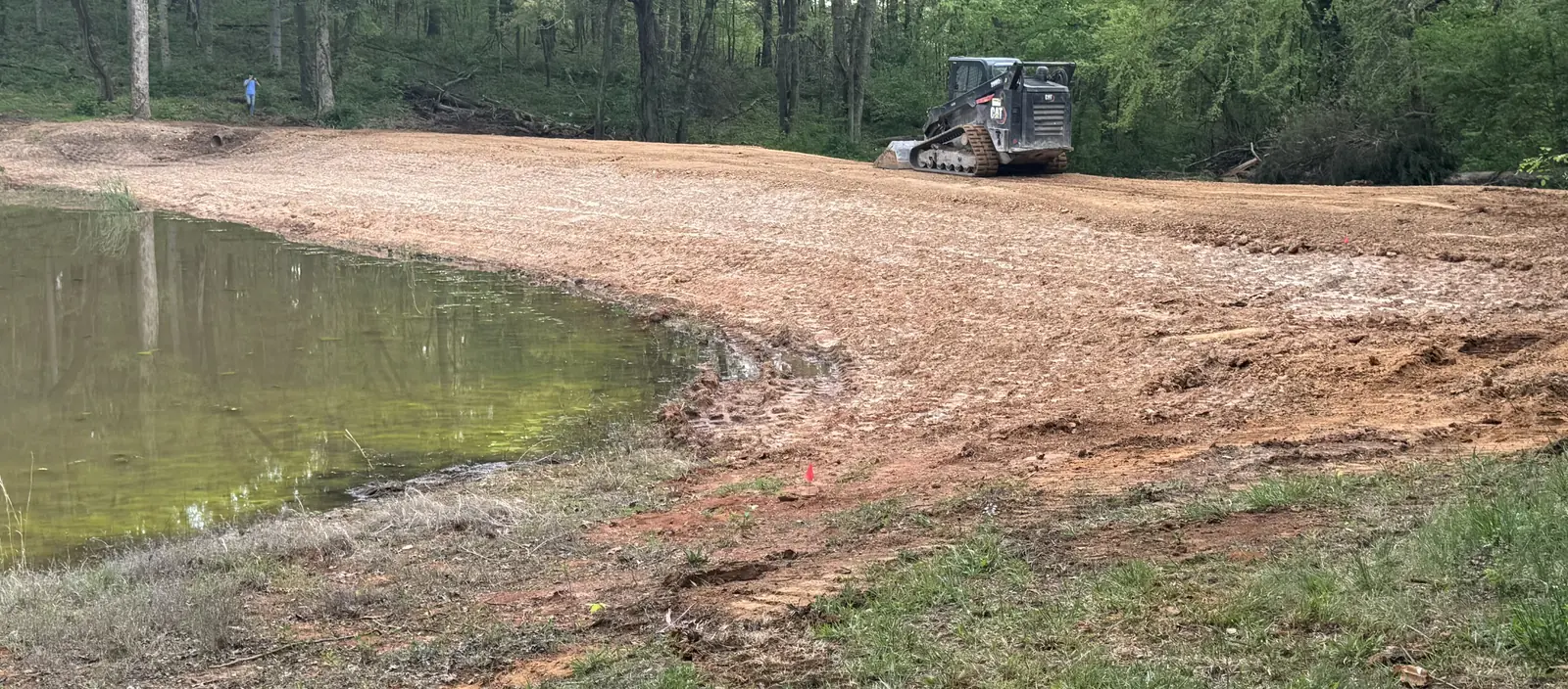 Repaired pond dam regraded and covered in straw, the compact track loader on top and the pond at the left
