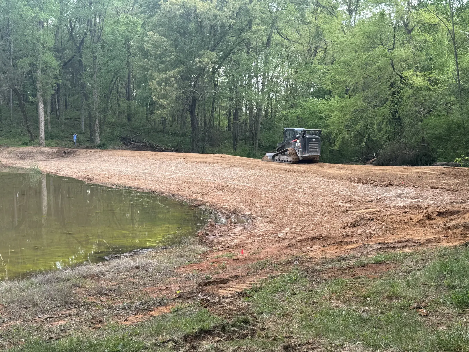 Repaired pond dam regraded and covered in straw, the compact track loader on top and the pond at the left